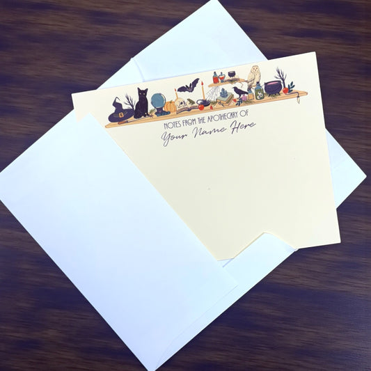 Set of flat cream notecards with witchy apothecary illustration and customizable name, paired with white envelopes on dark.