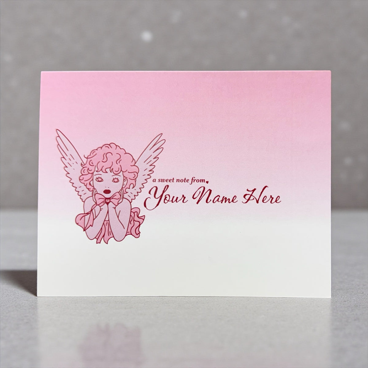 A2 folded note card with pink gradient background featuring a red sketch of a praying angel and customizable text "Your Name .