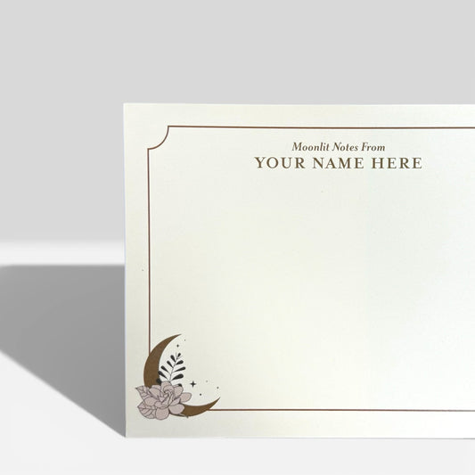 White flat note card with gold border, customizable name area, crescent moon, and floral design in bottom left corner.