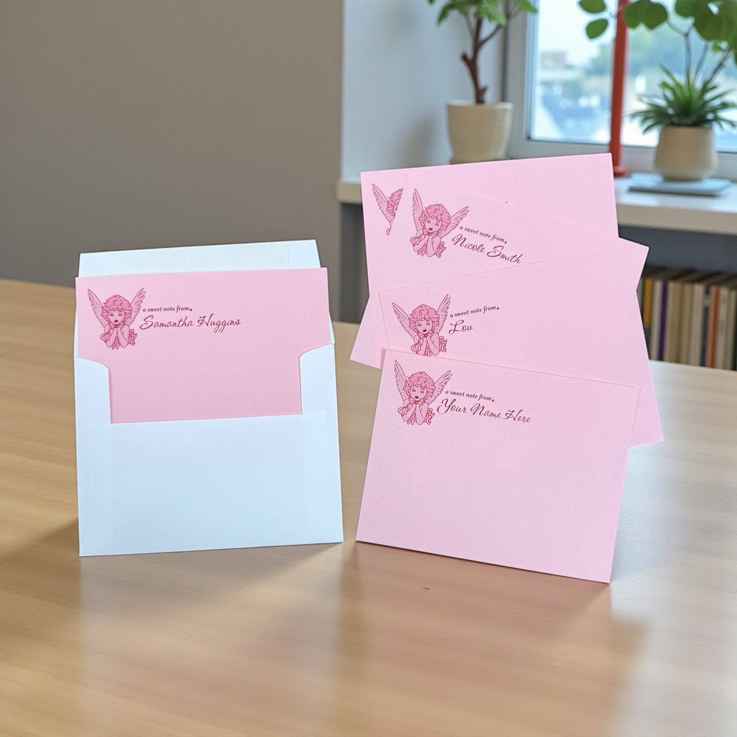 Set of pink personalized angel notecards with floral design and matching envelope on wood table.