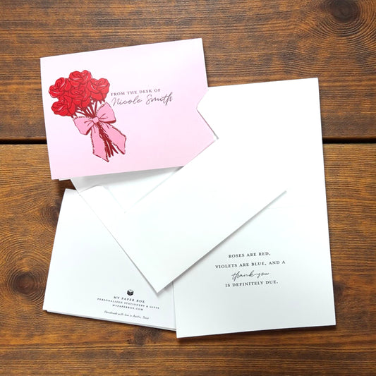 Pink card with a bouquet of red roses illustration and cursive text, shown with matching white envelopes and open card with.
