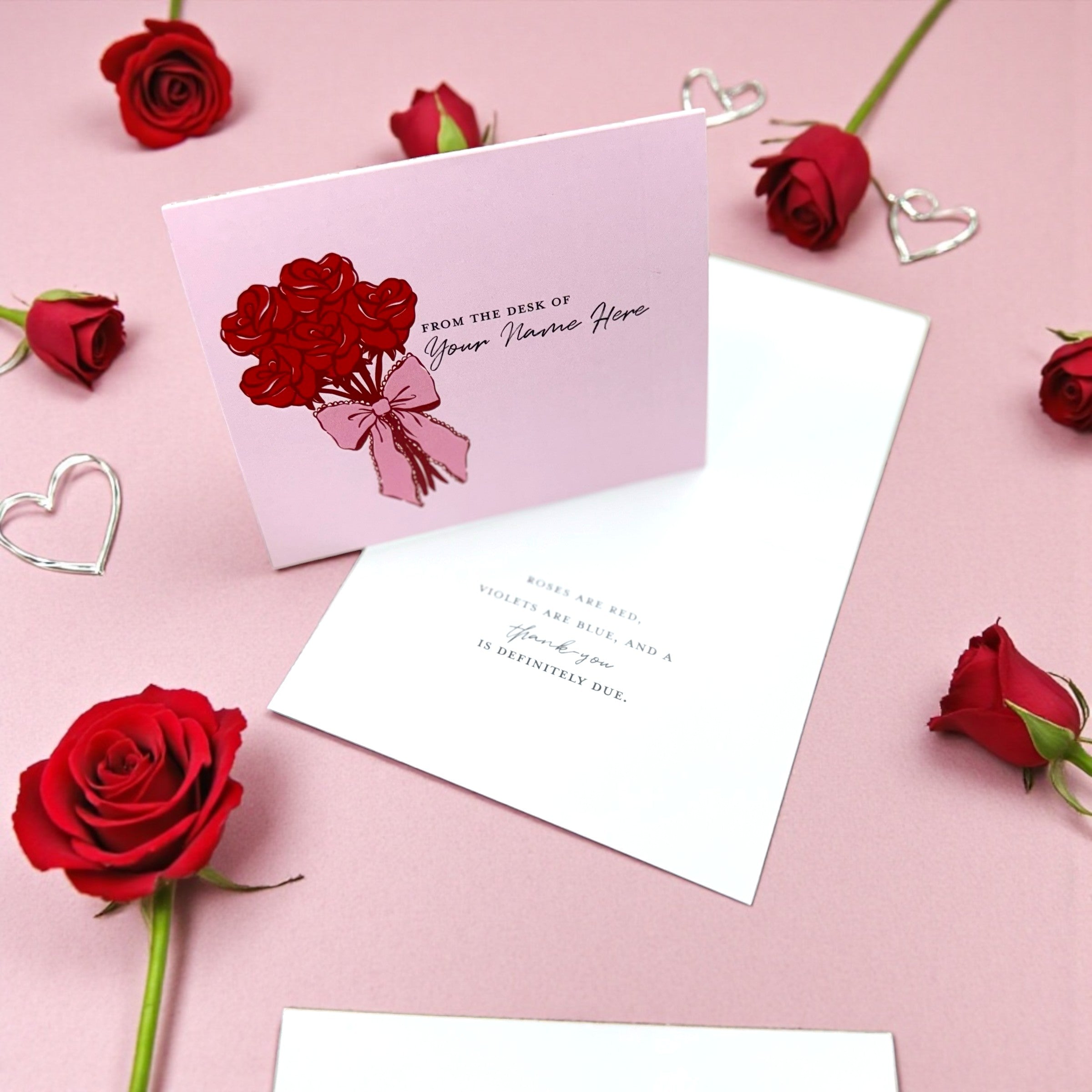 Pink greeting card with red rose bouquet illustration and handwritten-style text, surrounded by real red roses and silver.