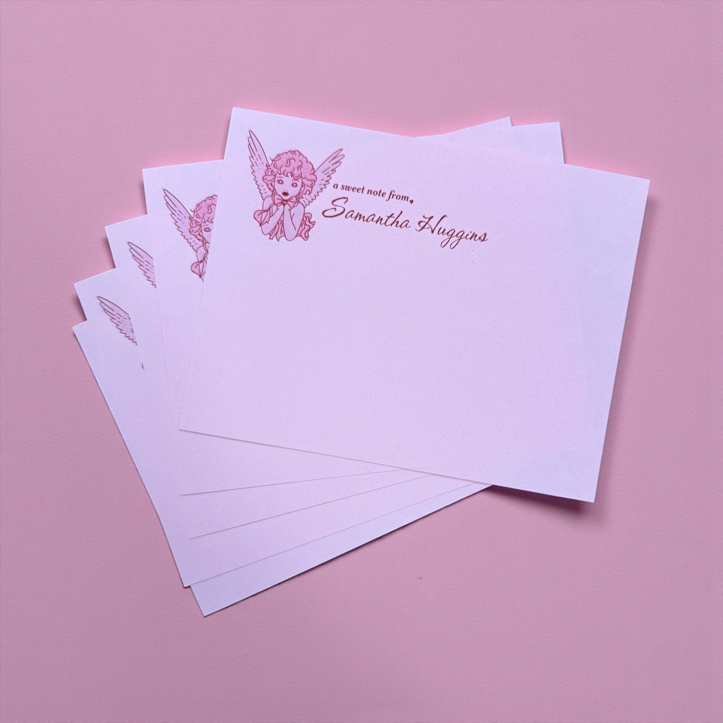 Stack of personalized stationery with angel design and text on a pink background