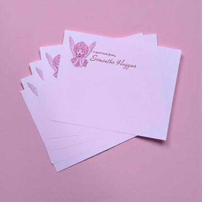Stack of personalized stationery with angel design and text on a pink background